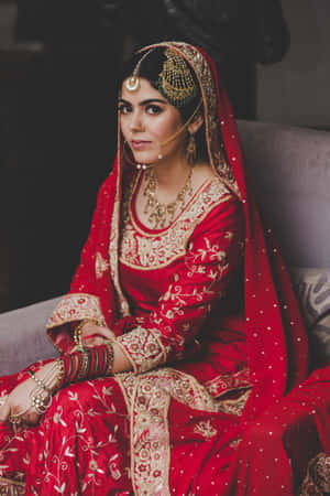 一位身穿紅金刺繡 lehenga 並佩戴珠寶的新娘坐在沙發上。 — 紅色 壁紙