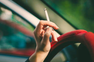 手在車內拿著點燃的香煙，紅色軚盤和模糊的綠色背景。