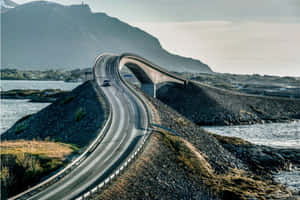 一座彎曲的現代橋樑橫跨水面，一輛汽車在靠近山脈的蜿蜒道路上行駛。 — 道路 壁紙