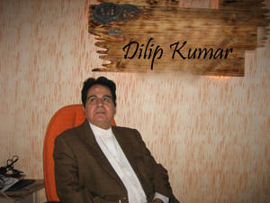 身穿棕色西裝的男子坐在橙色椅子上，上方有寫著 Dilip Kumar 名字的木牌。 — 橙色美學 壁紙