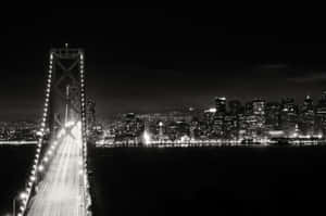 夜晚燈火通明的吊橋，背景是閃爍的城市天際線。 — 黑白 壁紙