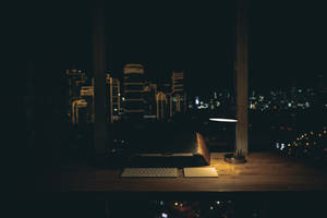 一張書桌，放著手提電腦和燈，在夜晚的城市天際線下被照亮。