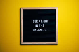 一個帶框的字母板，上面有訊息「I SEE A LIGHT IN THE DARKNESS」在黃色牆上。