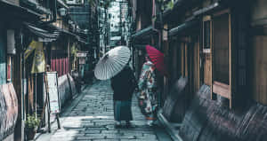 兩個穿著傳統日本服裝的人，沿著一條有木建築的狹窄石巷走著。