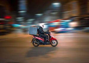 送貨員騎著紅色電單車，在模糊背景的城市夜晚街道上疾馳。 — 機車 壁紙