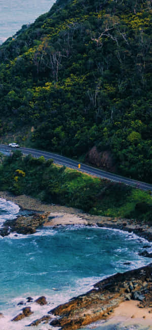 海岸公路蜿蜒穿過綠色山丘，旁邊是碧綠色的海洋和拍岸的波浪。 — iPhone 16 壁紙