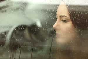 女性透過帶有雨痕的車窗向外看，表情陰鬱。 — 雨 壁紙