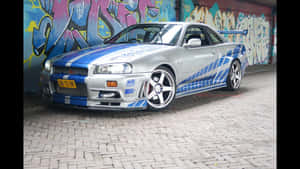 銀色日產Skyline R34帶藍色條紋停泊在塗鴉牆附近的磚街上。