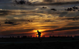 夕陽下田野上跑者的剪影，伴隨著戲劇性的橙色雲彩。 — 跑步 壁紙