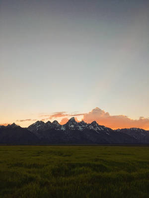 日落時分，白雪覆蓋的山峰在綠色草地後方升起。 — 簡約 iPhone 壁紙