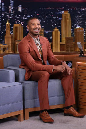 Michael B Jordan 坐在脫口秀佈景中微笑，身後有城市燈光。 — Jordan 標誌 GIF 壁紙