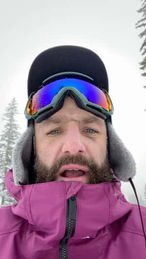 留鬍鬚的男子戴著滑雪護目鏡和紫色外套站在雪地森林中。 — 冬天 壁紙
