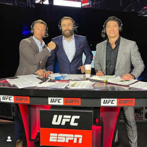 三位男性UFC ESPN評論員在廣播桌前，桌上放有文件和咖啡。