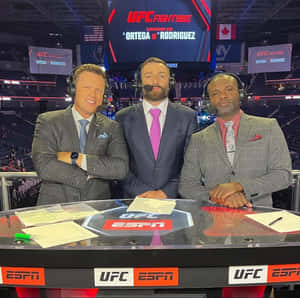 三位身穿西裝的男性UFC評論員在體育場館的ESPN轉播桌旁。