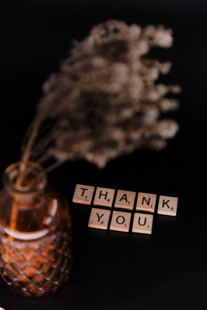 拼出 thank you 的 Scrabble 字母塊放在一個裝飾性琥珀色花瓶旁邊，花瓶裡有乾花。