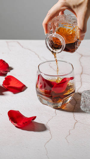 手在大理石上將琥珀色飲料倒入帶有玫瑰花瓣和冰塊的玻璃杯中。 — 玫瑰 壁紙