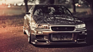 銀色日產Skyline R34在黃金時刻停泊在碎石路上，背景有樹木和城市燈光。
