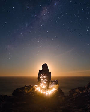 穿連帽衫的人夜晚坐在懸崖上，有串燈，觀看海洋上空的星星