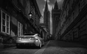 銀色Aston Martin停泊在狹窄的鵝卵石街道上，背景有哥德式塔樓。 — 黑白 壁紙