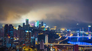 多雲天空下，河流上有燈光建築和橋樑的夜晚城市。 — 夜景城市 壁紙