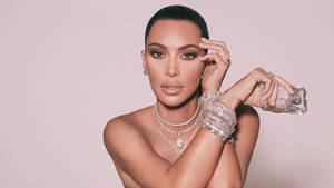 Kim Kardashian 手持香水瓶，佩戴閃亮珠寶，背景為粉紅色。