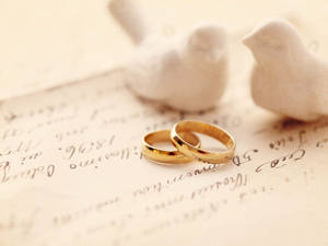 兩枚金色結婚戒指放在舊手寫信上，旁邊有白色小鳥雕像。 — 白鴿 壁紙