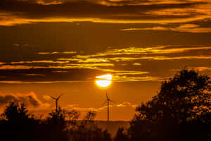 風力渦輪機高聳於鮮豔的橙色夕陽天空，伴隨著深色樹木的剪影。 — MacBook Pro 壁紙