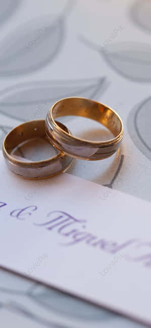 兩隻金色結婚戒指放在帶有名字和葉子設計的婚禮邀請函上。 — 銀河A 壁紙