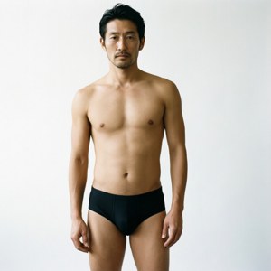 一個肌肉發達的男人穿著黑色內褲站在白色攝影棚中。