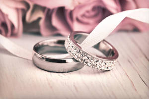 兩枚結婚戒指，一枚素面，一枚鑲鑽，放在白色絲帶上，配以粉紅玫瑰。 — 3:2 壁紙