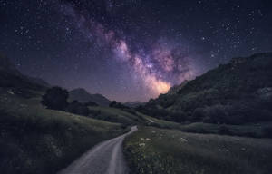 星空之夜，銀河高懸於山巒景觀之上，一條泥土小徑蜿蜒穿過草地。