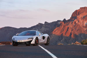 銀色McLaren GT6 MCL跑車在沙漠公路上，日落時分紅色山脈。
