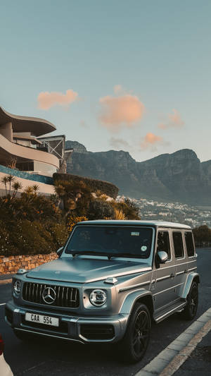 銀色 Mercedes G-Wagen 停泊在現代建築附近，背景是山脈和日落。