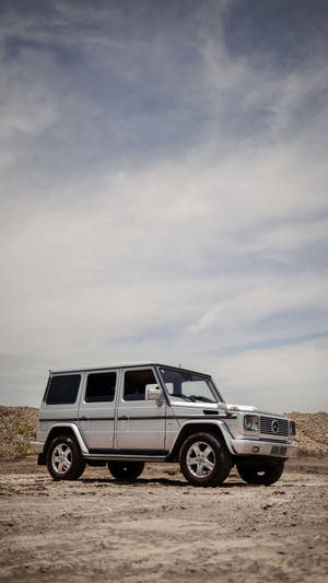 銀色 Mercedes-Benz G-Class SUV 停喺乾燥嘅泥地上，上面係多雲嘅天空。