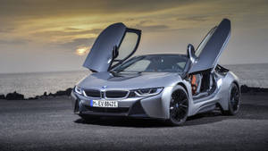 銀色BMW i8，鷗翼車門打開，停在日落時分的岩石海灘上。