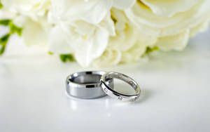 白色表面上白玫瑰旁的兩枚結婚戒指。 — 16:10 壁紙