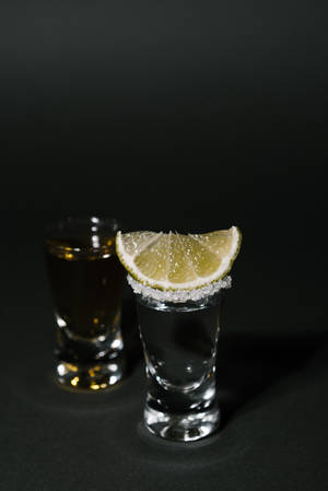 兩個烈酒杯，一個有青檸角和鹽邊，另一個深琥珀色在黑色表面上。