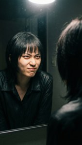 身穿黑色皮褸的人，在明亮燈光下對著鏡中倒影做出兇狠表情。