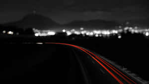 夜間高速公路上的紅色光軌，遠方有模糊的城市和山脈。 — 暗夜城市 壁紙