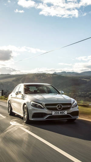 銀色 Mercedes 房車在風景優美的山路上快速行駛，背景有山丘和藍天。
