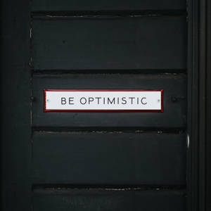 一個白色帶紅色邊框的標誌，上面寫著 BE OPTIMISTIC，位於深綠色木門上。