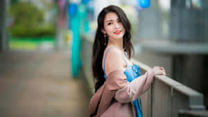 年輕女子穿著藍色連衣裙和粉紅色外套，微笑著靠在欄杆上。