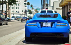 藍色Aston Martin DB9停泊在帶有棕櫚樹和建築物的城市街道上。