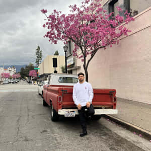 男人靠在復古紅色卡車旁，旁邊是粉紅色花樹，在城市街道上。