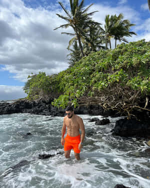 穿著橙色泳褲的男子在熔岩岩石和棕櫚樹附近的海浪中涉水。