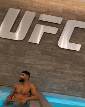 一個留鬍子的男人在木牆上一個大型UFC標誌下的藍色水池中放鬆。 — 健身 壁紙