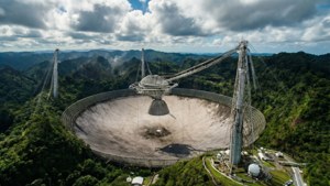 在多雲天空下，大型無線電望遠鏡碟形天線被茂密的綠色森林和山脈環繞
