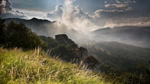 陽光穿過雲層，照耀在雲霧繚繞的山脈和綠色山谷上，有高高的草叢。 — 天際線 壁紙