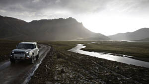 銀色 Land Rover Defender 在日落時分靠近河流的泥濘小路上，背景有山脈。