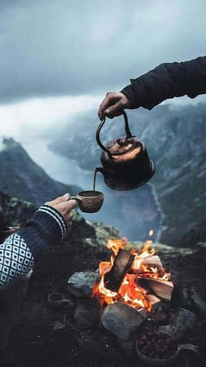 在崎嶇山地中，有人將水壺中的茶倒在營火上。 — 露營 壁紙